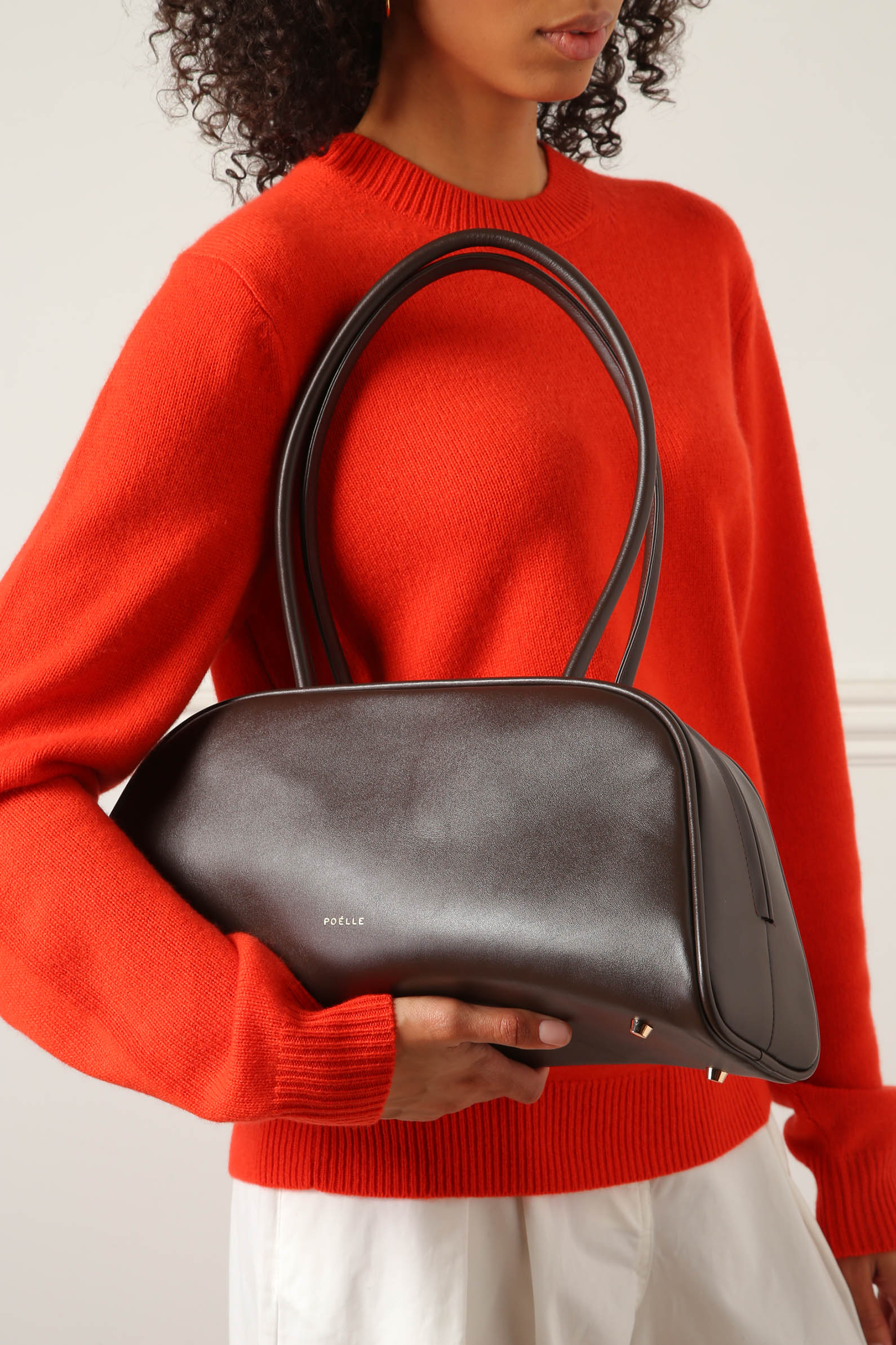Poelle Atelier east-west horizontal leather bag in dark brown held by model wearing red sweater