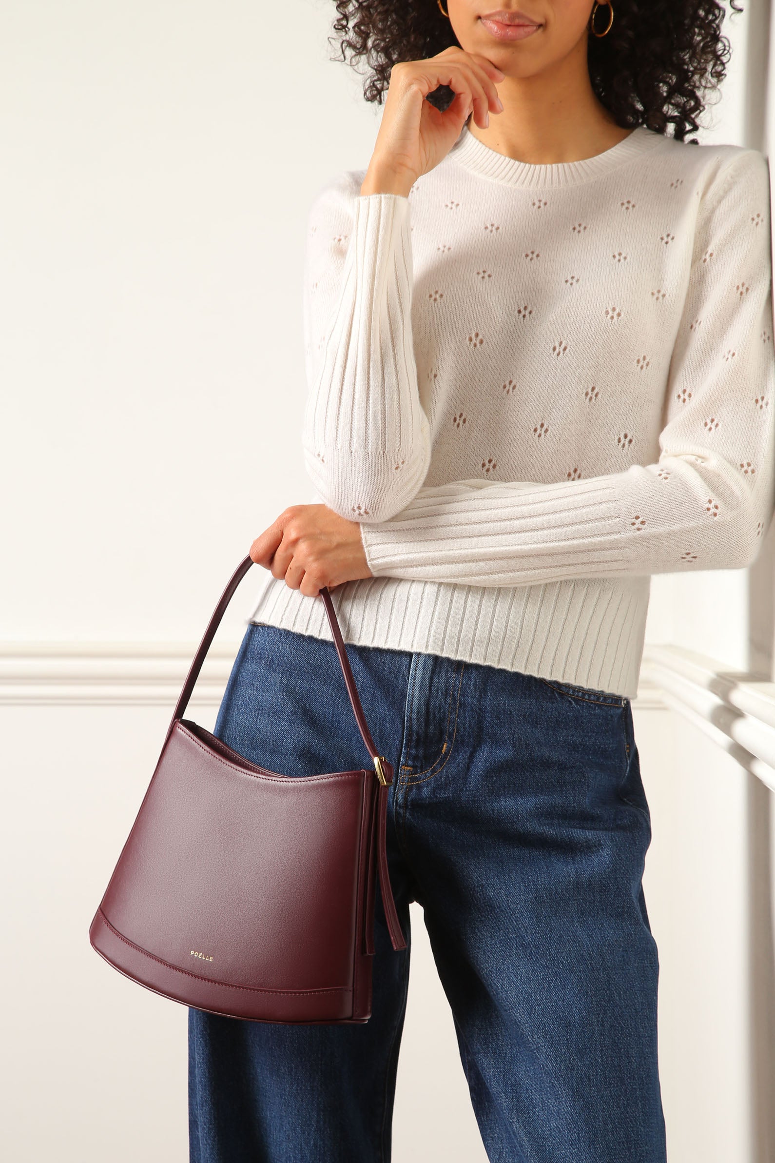 Poelle Atelier luxury minimalist leather bag in burgundy held by a woman wearing white sweater and blue jeans